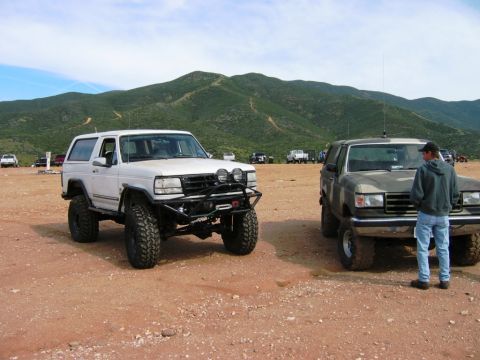 1985 Ford Bronco - Two modified Ford Broncos are parked off-road with hills in the background.