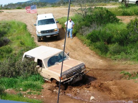1985 Ford Bronco - A tan Ford Bronco is stuck in a muddy ditch while a white Ford SUV waits behind it on a dirt path.