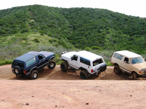 1985 Ford Bronco - Three Ford Broncos, a white, tan, and blue model, navigate a dusty trail with a green, hilly landscape in the background.