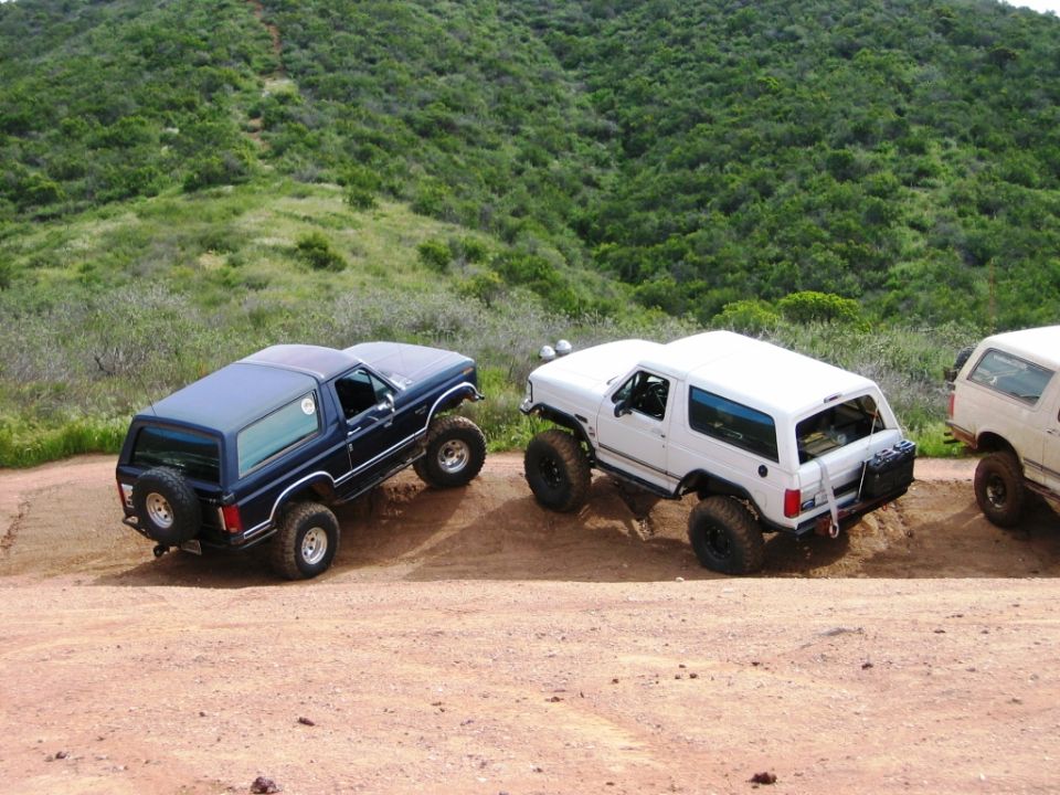 Blue and white lifted Ford Broncos on dirt mound off-road, green hills behind