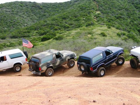 1985 Ford Bronco - Several Ford Bronco SUVs are lined up on a dirt path, with a grassy hillside behind them.