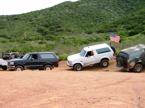1985 Ford Bronco - Multiple Ford Broncos navigate a dusty trail, with one flying the American flag.