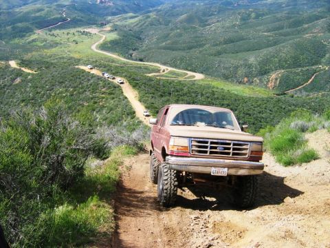1985 Ford Bronco - A red Ford Bronco navigates a winding dirt road through a mountainous landscape with other vehicles in the distance.