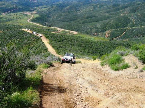 1985 Ford Bronco - Several off-road vehicles navigate a challenging dirt path through a hilly, green landscape.