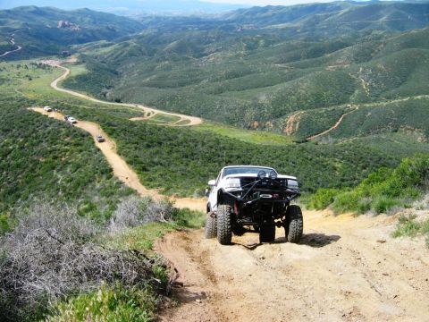1985 Ford Bronco - Several vehicles navigate a winding dirt road through a hilly, green landscape.