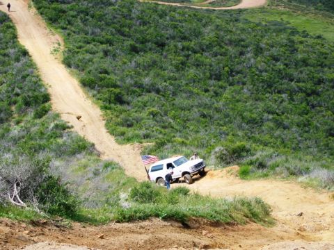 1985 Ford Bronco - A white SUV navigates a steep dirt path, an American flag waving from its rear.