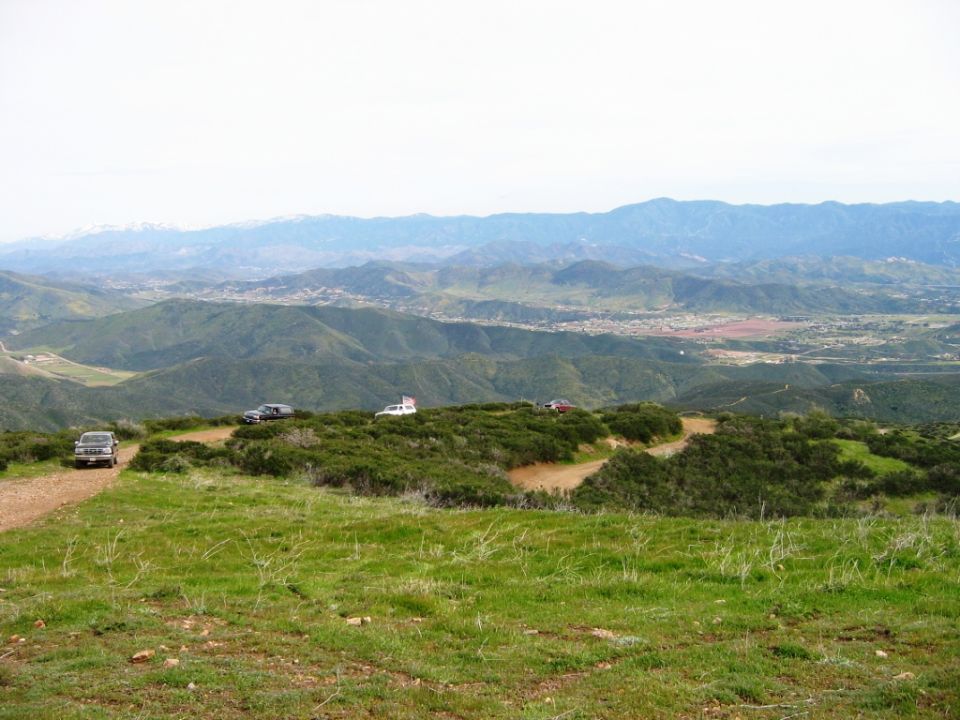 Vehicles parked on a grassy hilltop dirt trail overlooking green hills and distant mountains