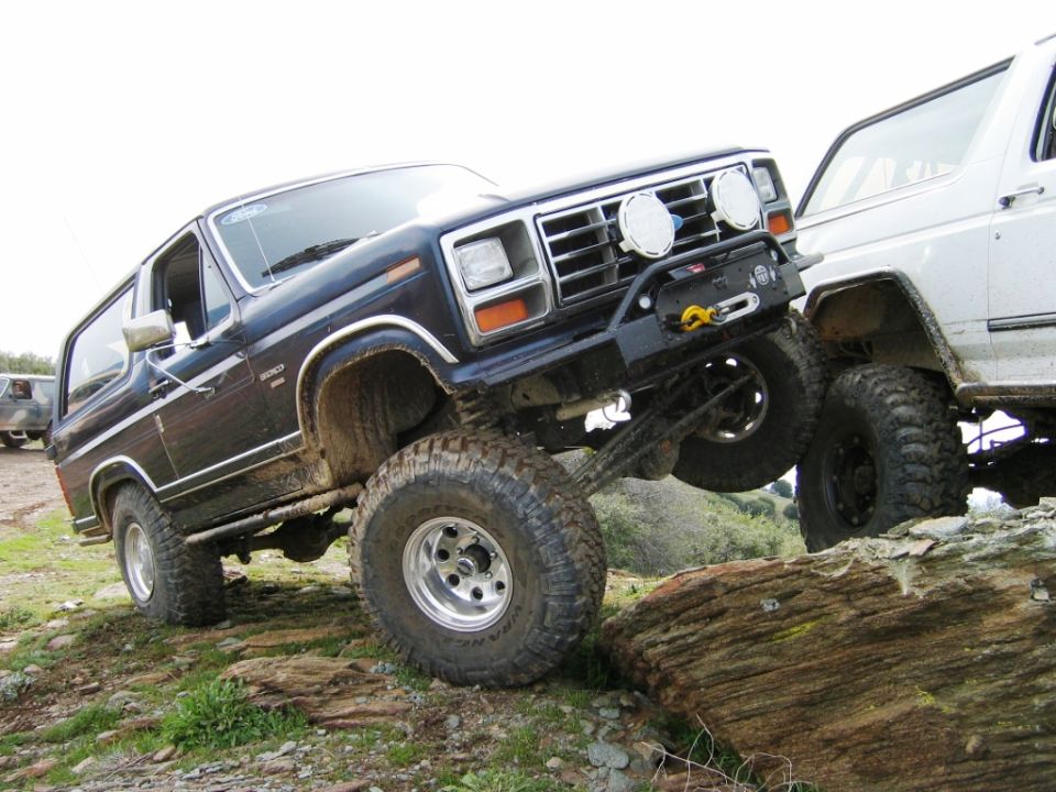 Dark Ford Bronco flexing over a log obstacle off-road with mud tires and brush guard