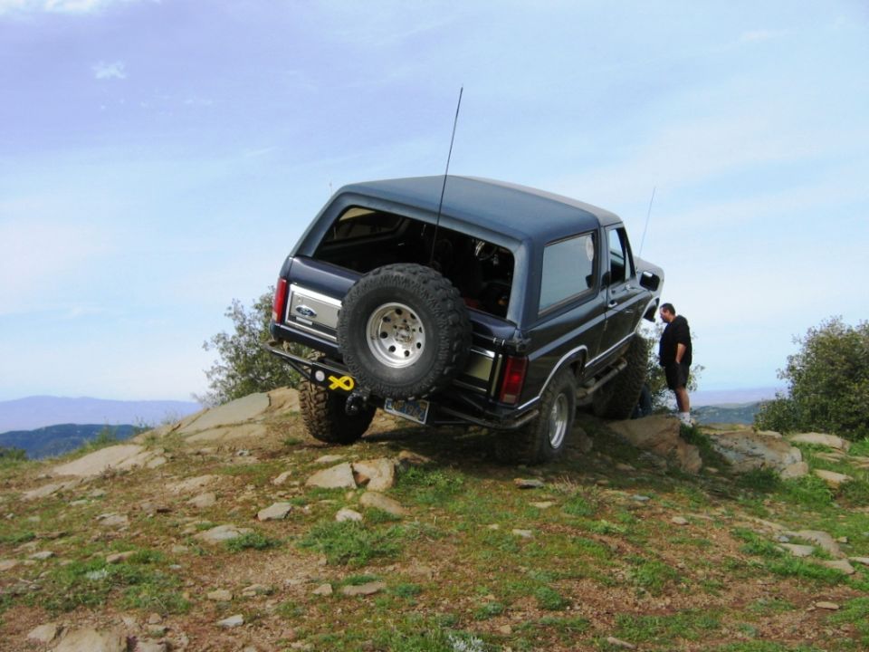 Dark Ford Bronco SUV rear view climbing rocks, man standing beside it on a hill