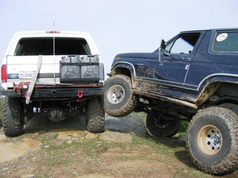 1985 Ford Bronco - Two Ford Broncos, one white and one blue, are shown off-roading on a rocky terrain.