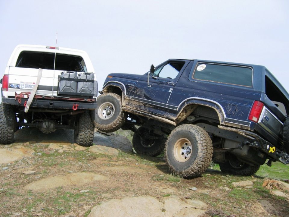 Dark blue lifted Ford Bronco on mud tires climbing rocks beside a white Ford truck