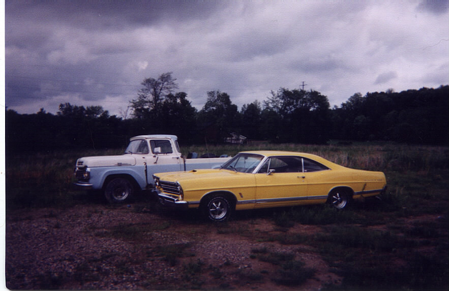 Yellow 1967 Ford Galaxie parked next to a blue and white pickup truck under a cloudy sky