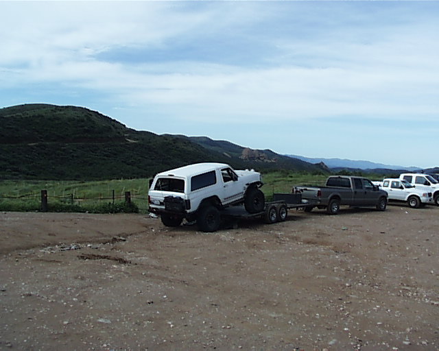 white SUV loaded onto a trailer being towed by a pickup truck, with other trucks parked nearby
