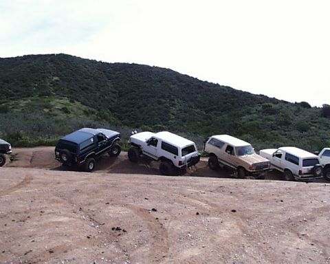 1993 Ford Bronco - A group of Ford Bronco SUVs are lined up on a dirt road, with a grassy hill in the background.