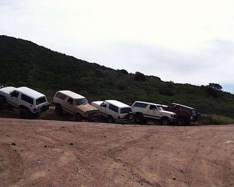 1993 Ford Bronco - Several Ford Bronco SUVs are parked on a dirt road with a hill and cloudy sky behind them.