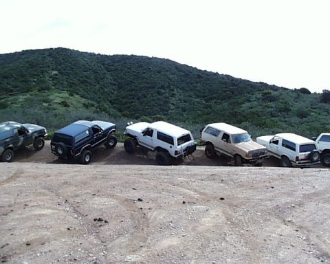 1993 Ford Bronco - A line of Ford Bronco SUVs are parked on a dirt road, with a grassy hill behind them.