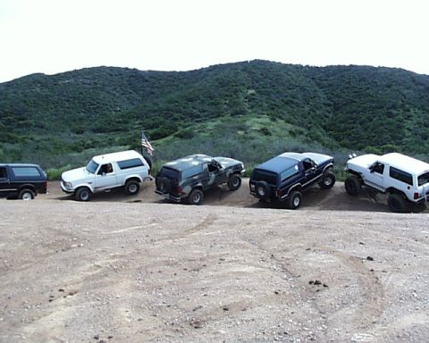 1993 Ford Bronco - A lineup of Ford Broncos is parked on a dirt road, with a flag flying in the background.