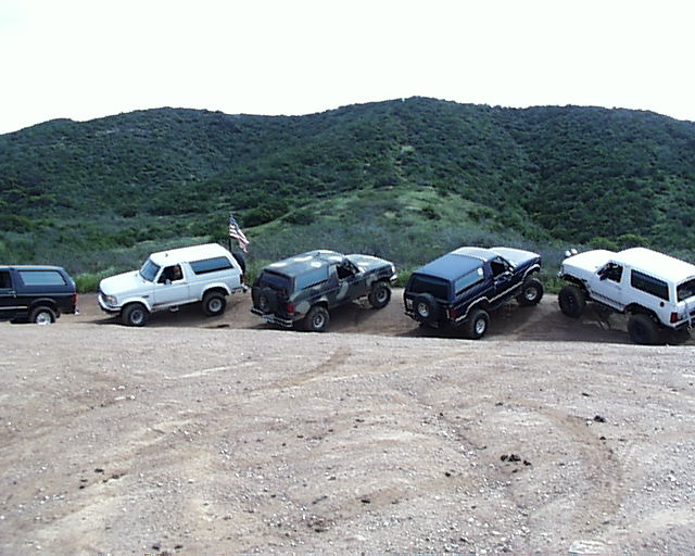 Five Ford Broncos parked on a dirt road with a hilly, green background. An American flag is visible