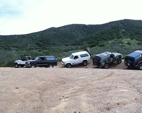 1993 Ford Bronco - Several SUVs, including a dark blue Ford Bronco, are parked on a dirt road with a hill in the background.