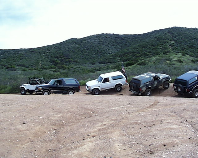 Four-door Ford Bronco parked on a dirt road, with a white Jeep and two other SUVs behind it