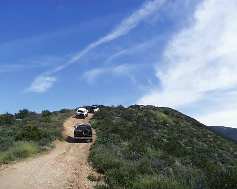 1993 Ford Bronco - Multiple SUVs navigate a winding dirt path up a grassy hill under a bright blue sky with scattered clouds.