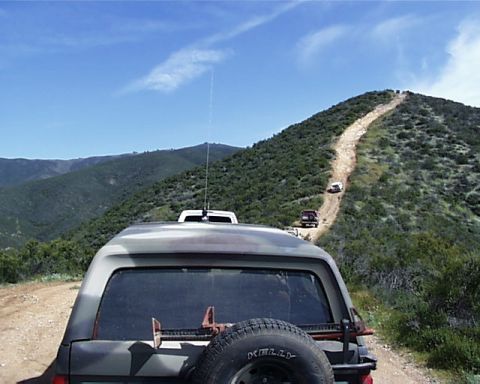 1993 Ford Bronco - Several SUVs ascend a steep, winding dirt road through a green, hilly landscape under a clear blue sky.