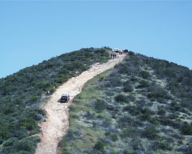 Dark SUV climbing a steep rocky dirt trail up a brushy hill, with people and another vehicle near the top.