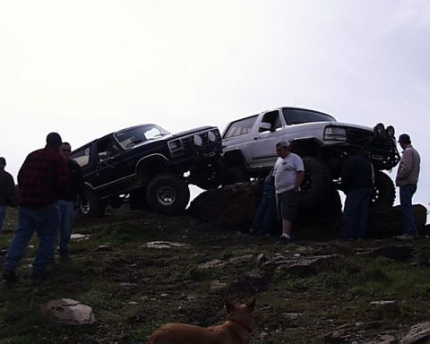 1993 Ford Bronco - Two lifted Ford Broncos navigate a rocky off-road course with onlookers present.