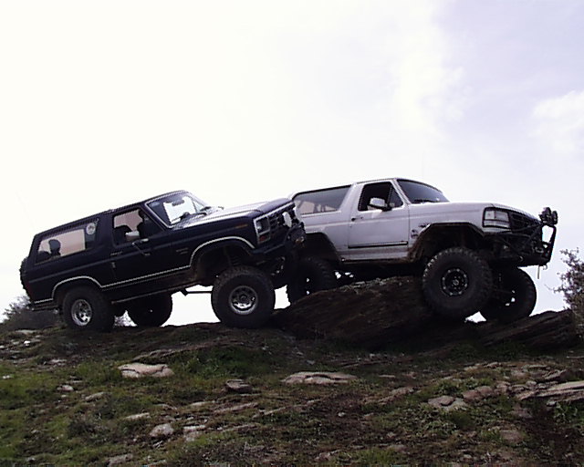 Dark and white lifted Ford Broncos parked nose-to-nose on rocks against a cloudy sky