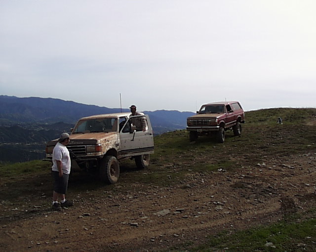 White Ford Bronco and red Ford Bronco on a dirt hill with mountains in the background