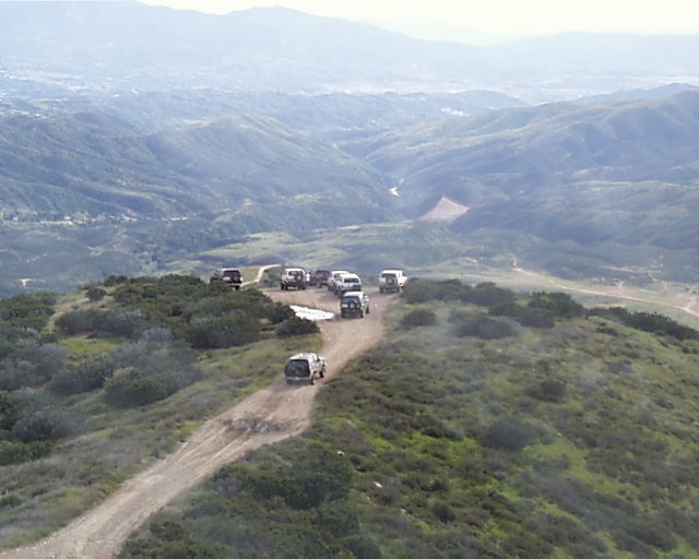 Several SUVs drive on a dusty mountain road, with a town visible in the distant valley