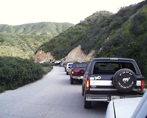 1993 Ford Bronco - A convoy of SUVs drives down a winding road through a hilly, vegetated landscape.