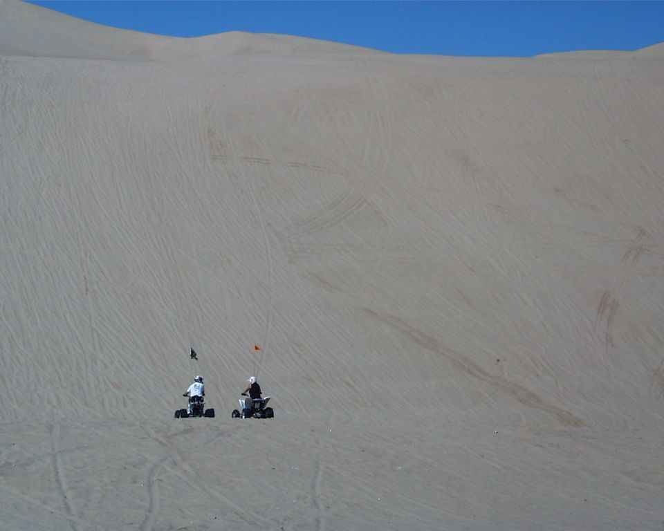 Two ATVs ride on a vast sand dune under a clear blue sky