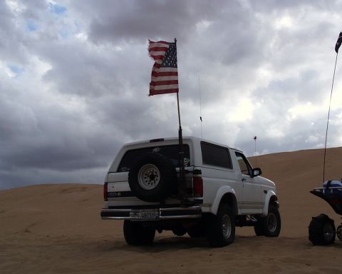 1993 Ford Bronco - A white 1993 Ford Bronco is parked on a sandy dune under a cloudy sky, with an American flag attached to the back.