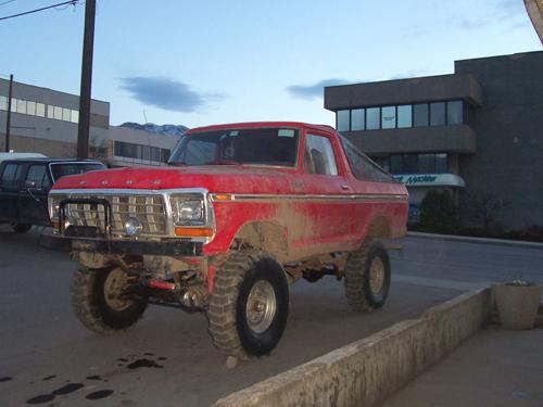 Lifted red Ford Bronco with large mud tires and dirty body parked on street near buildings