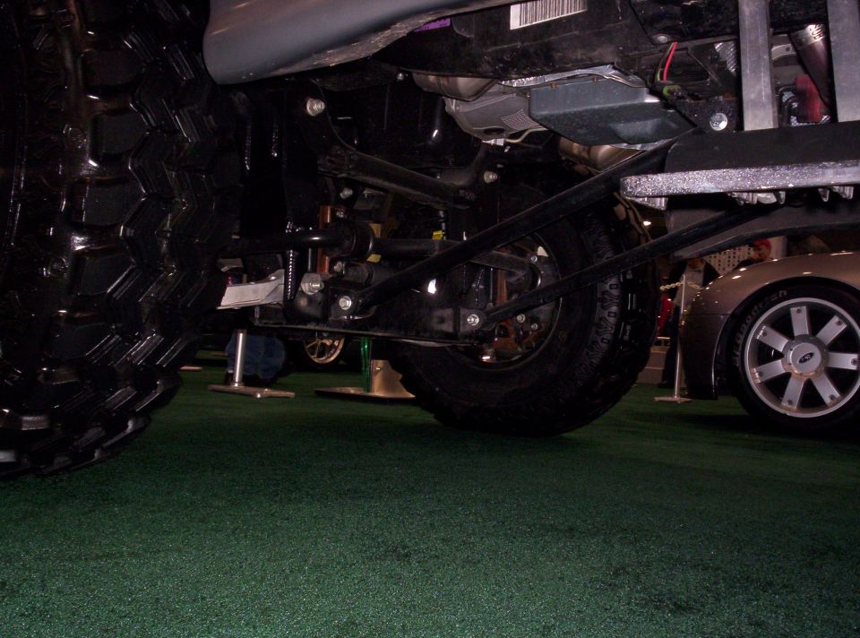 Underside of a large vehicle with off-road tires, showing suspension and a silver car wheel with a Ford logo