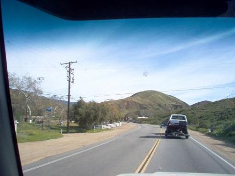 1988 Ford Bronco - A white pickup truck tows a vehicle on a trailer along a rural highway with rolling hills in the background.