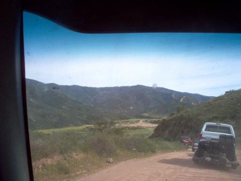 1988 Ford Bronco - Several vehicles travel on a dirt road through a hilly, green landscape under a bright sky.