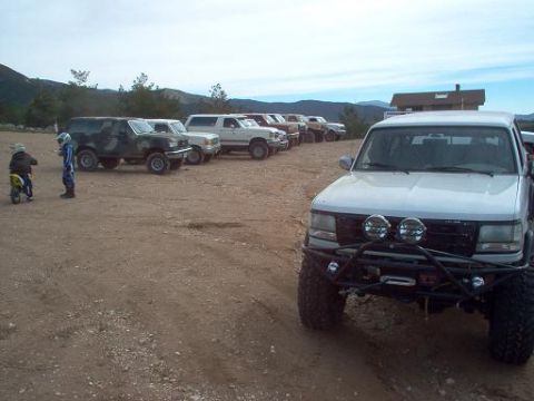 1988 Ford Bronco - Several off-road vehicles are parked in a dirt lot with mountains in the background.