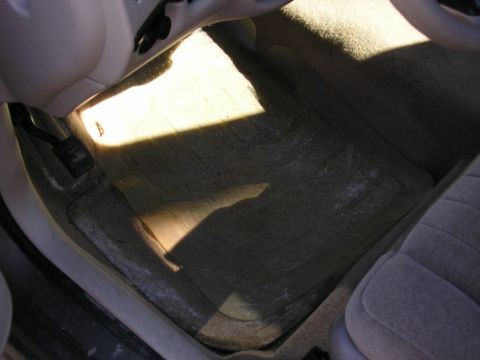 1998 Ford Taurus - The interior floor of a car shows dirt and a shadow from sunlight, with pedals and a seat visible.
