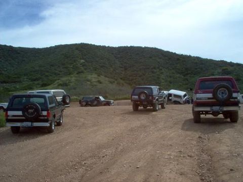 1988 Ford Bronco - Several SUVs are parked on a dirt road with a grassy hill in the background.