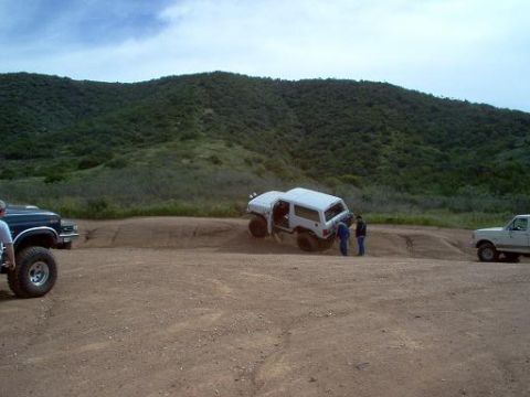 1988 Ford Bronco - A white 1988 Ford Bronco is seen tilted on a dirt trail with its doors open, while two individuals observe the scene.