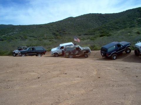 1988 Ford Bronco - A group of Ford Bronco SUVs are parked on a dusty trail, with a hilly landscape under a partly cloudy sky.