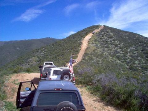 1988 Ford Bronco - Several off-road vehicles ascend a steep, dusty mountain trail under a clear blue sky.
