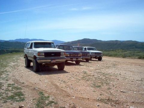 1988 Ford Bronco - Three Ford vehicles, including a Bronco and two pickup trucks, are parked on a dirt road with a mountainous landscape in the background.