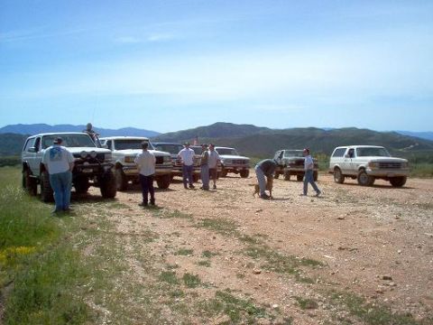 1988 Ford Bronco - Several Ford Broncos and F-Series trucks gathered on a dirt road with people and a dog in a scenic, hilly area.