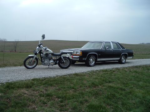 1989 Ford Crown Victoria LTD - A black sedan and a motorcycle are parked side-by-side on a gravel road under an overcast sky.