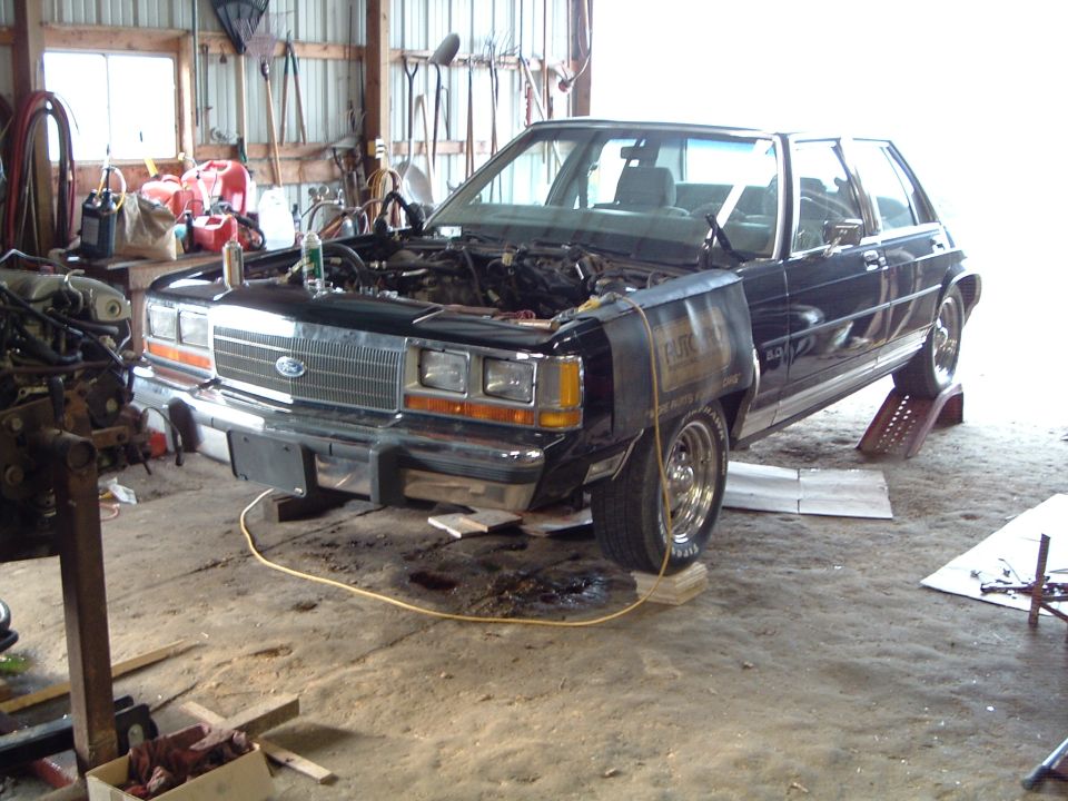 Dark Ford Crown Victoria with hood removed being worked on in a pole barn shop