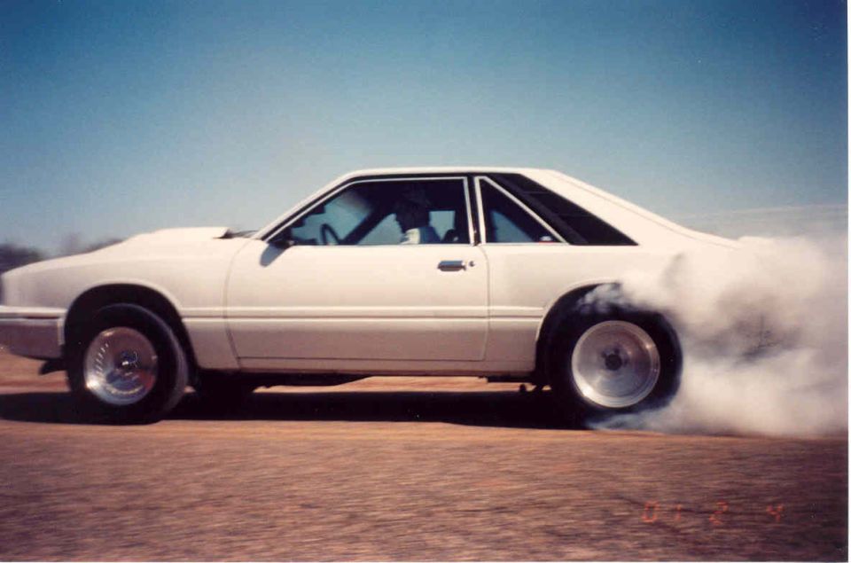 White Fox-body Mercury Capri coupe doing a smoky rear-wheel burnout on a dirt lot