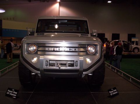 1989 Ford Bronco - Silver SUV with "BRONCO" lettering on the grille is displayed at an auto show, with other vehicles visible in the background.